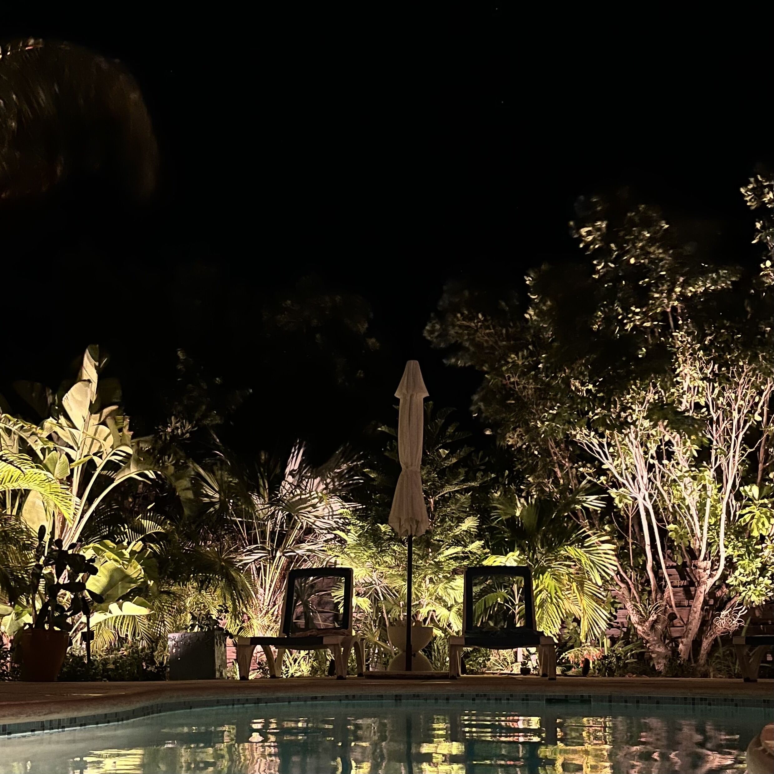 Two loungers and a closed umbrella stand at the pool at night.