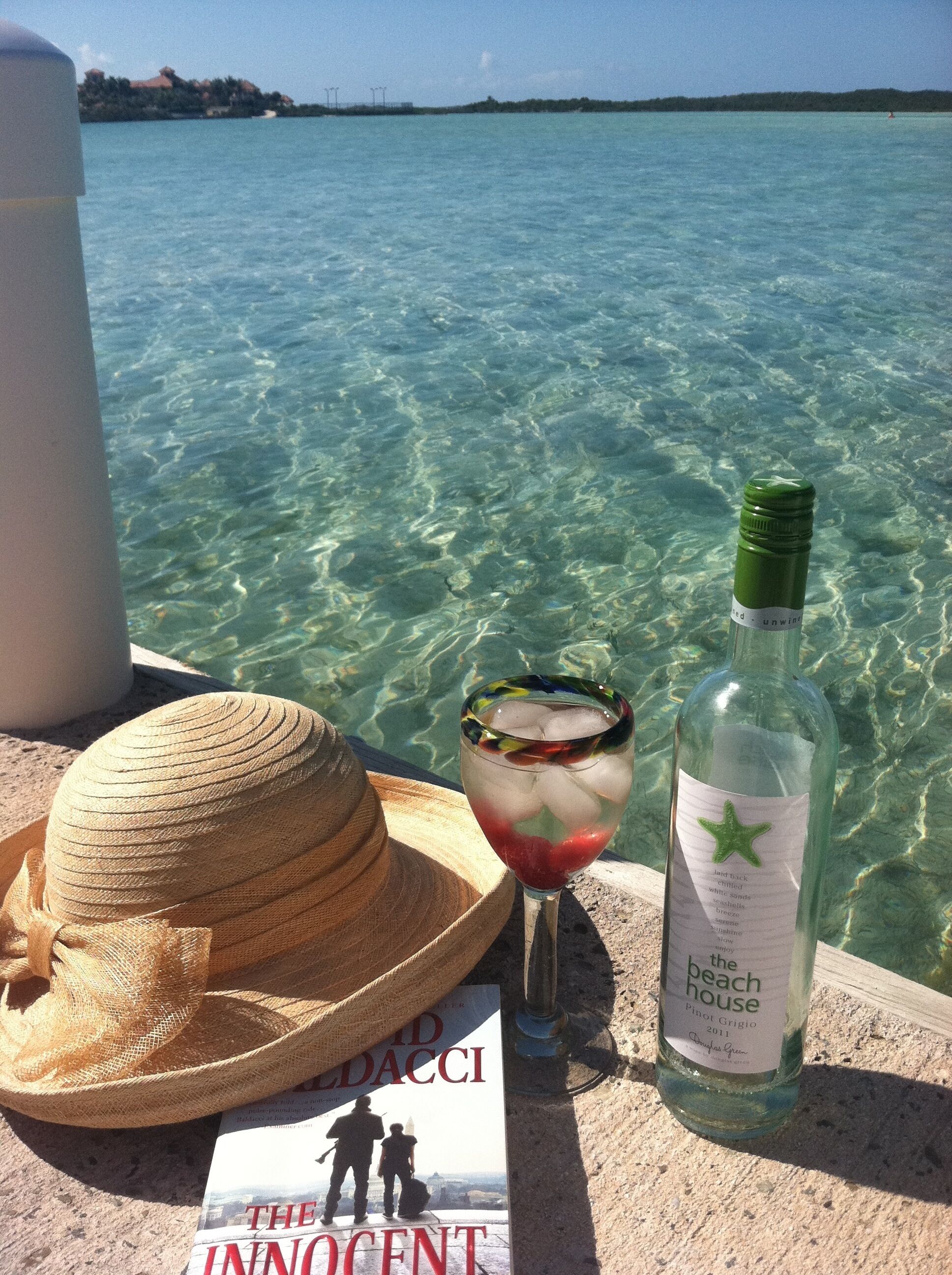 A sun hat, a glass, and a bottle on a wall next to clear water.
