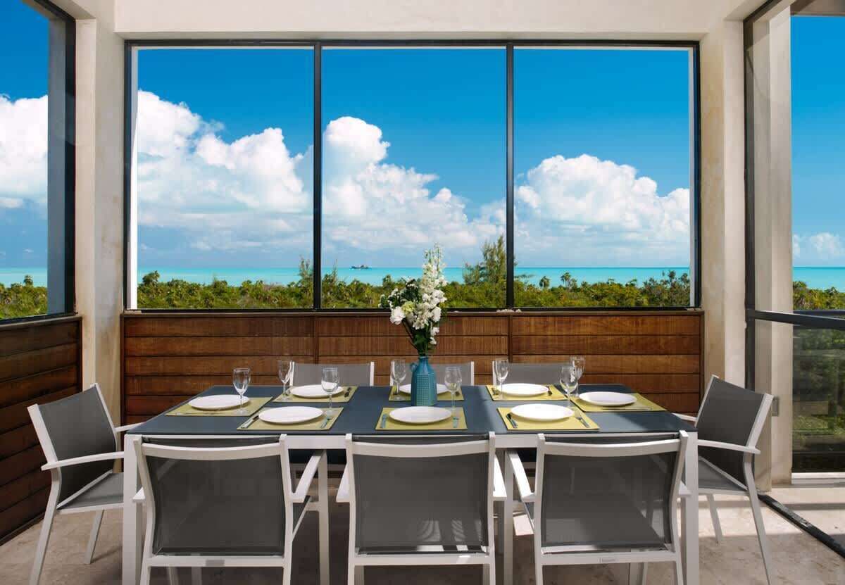 A dining table with eight chairs stands before large windows with a view of the sea.