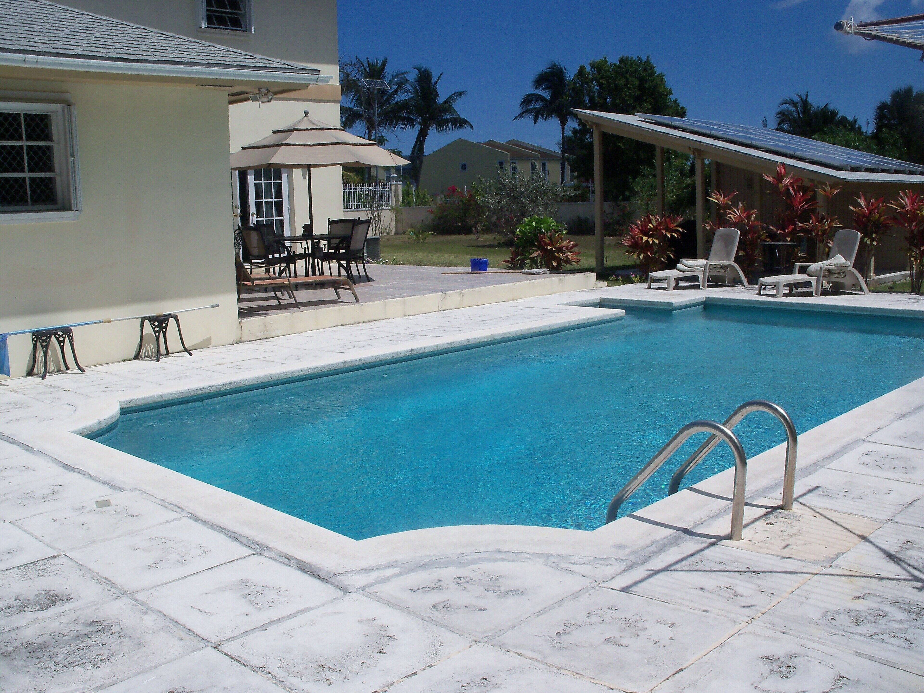 The pool has a stone deck, a patio with an umbrella, and palm trees behind the villa.