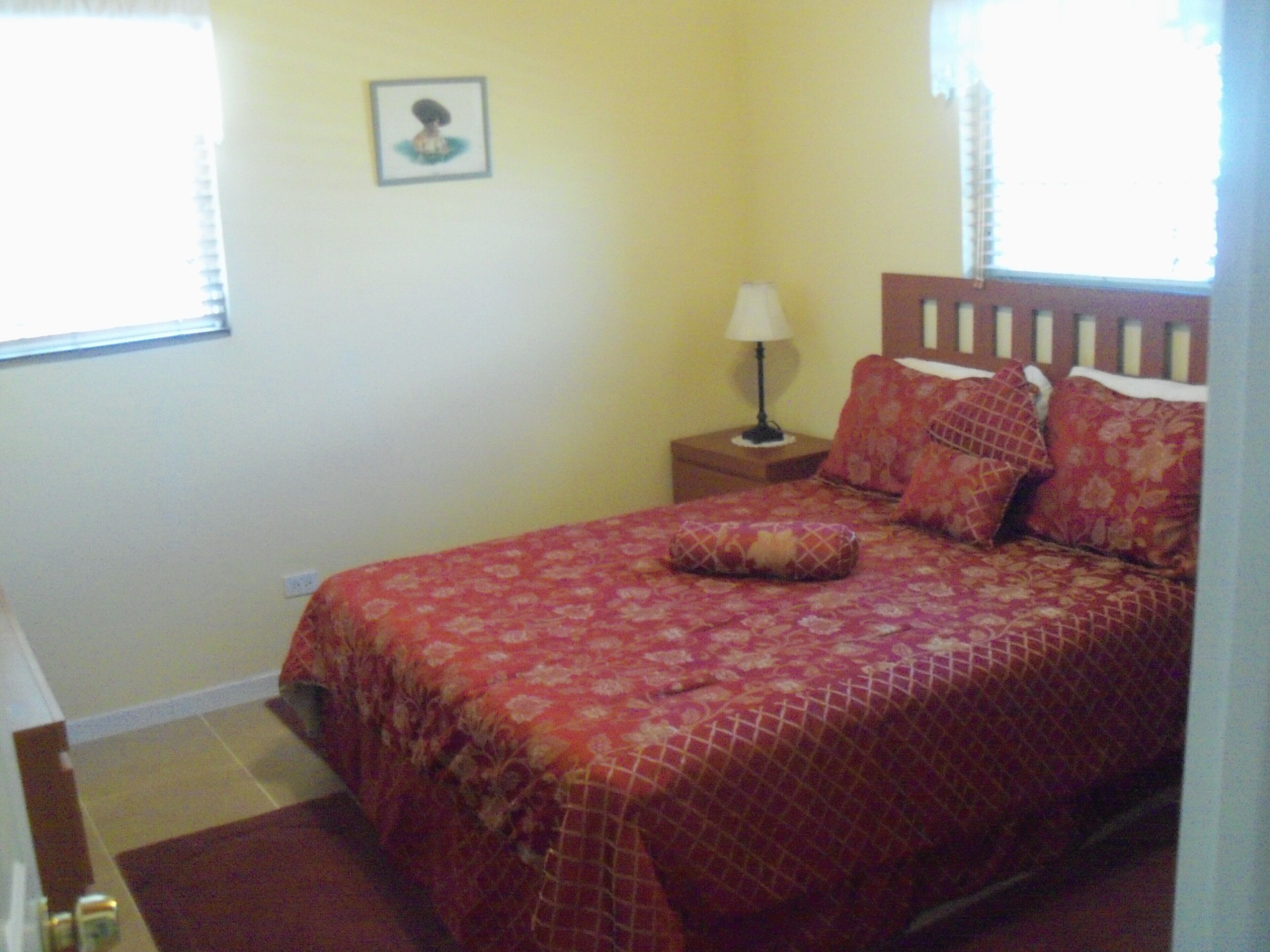 The bedroom has a double bed with a red cover and a nightstand with a lamp.