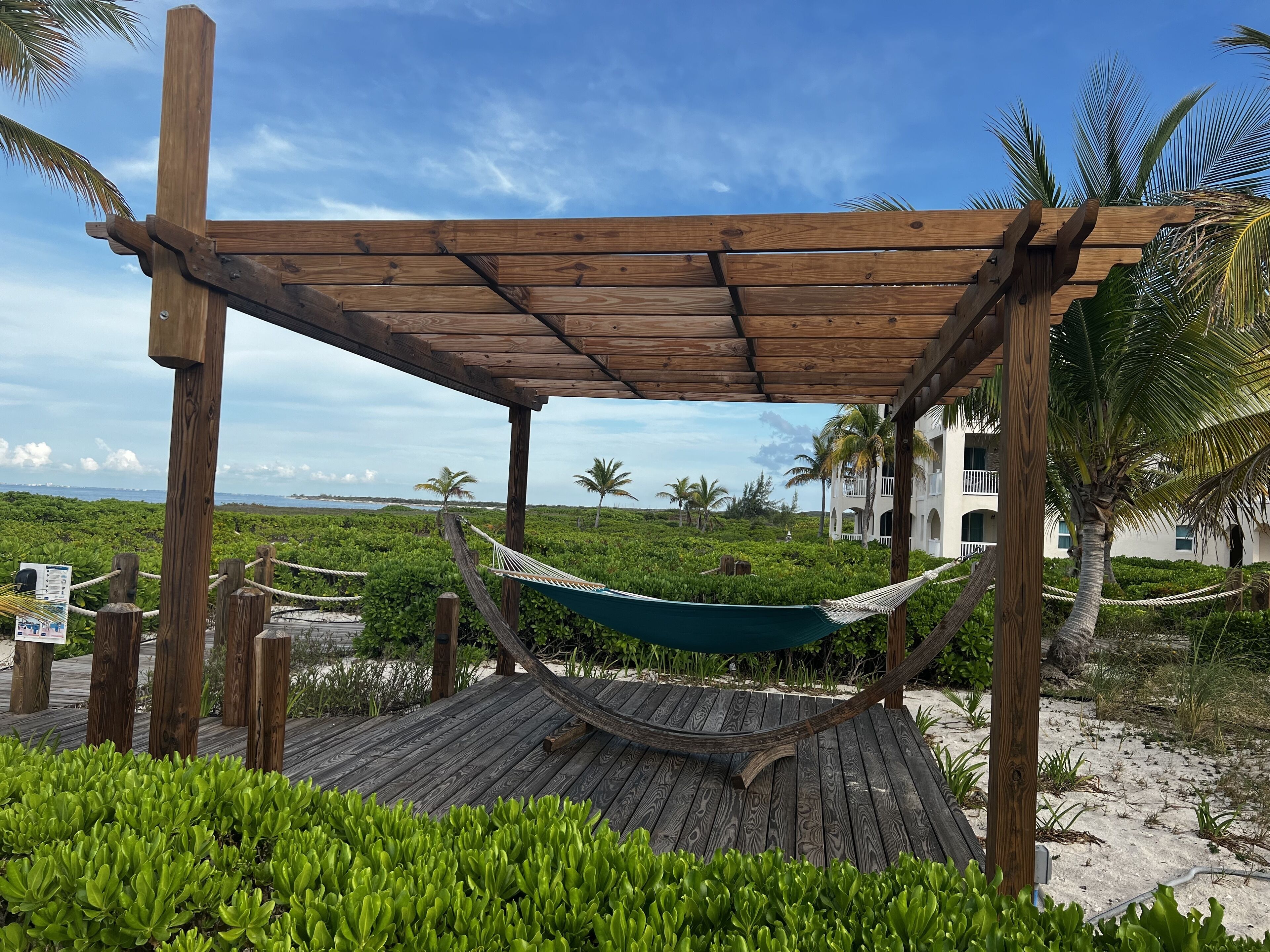 A hammock under a wood pergola on a deck next to the beach.