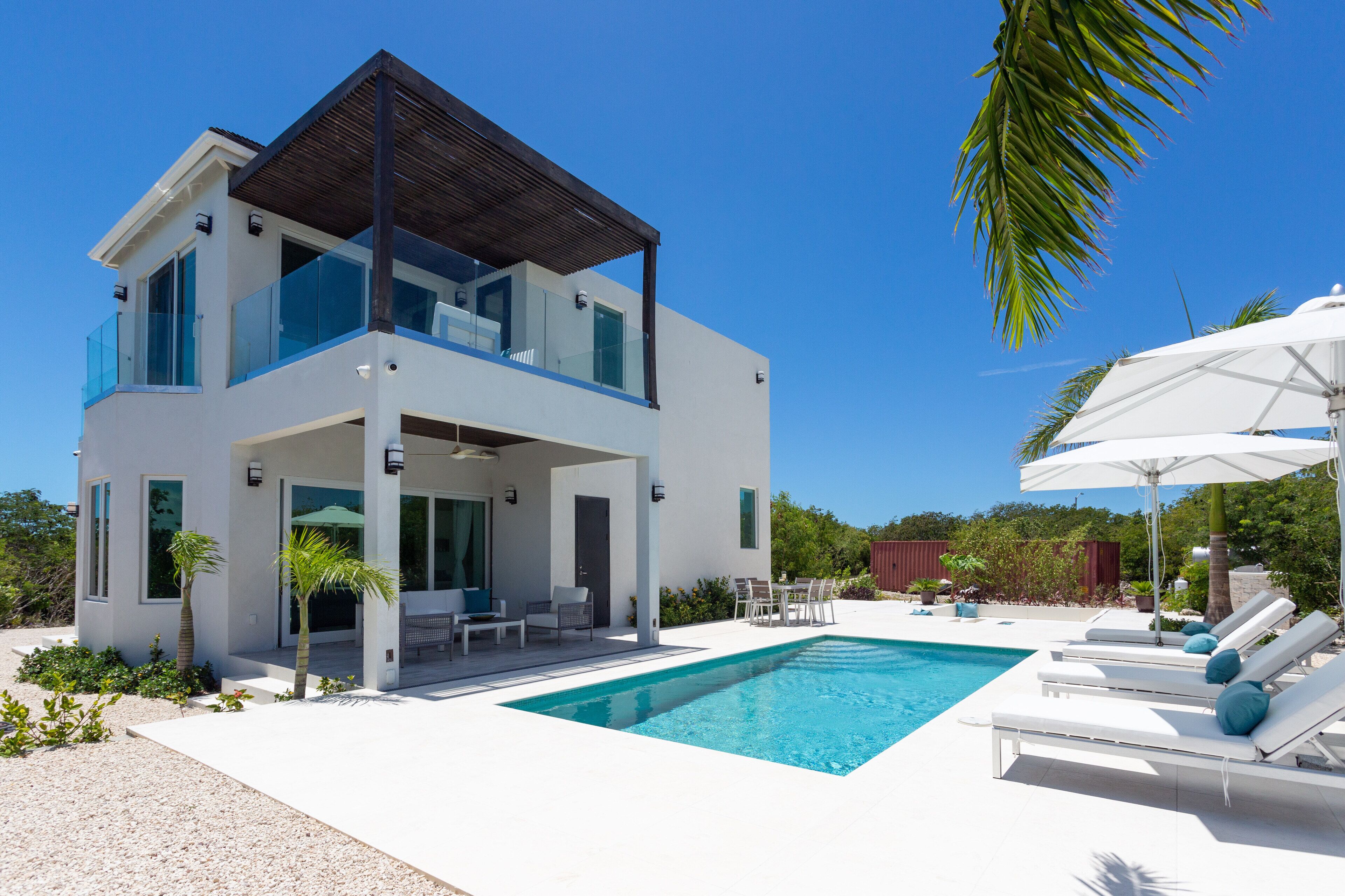 The two-story white villa with a balcony, the pool, and loungers.
