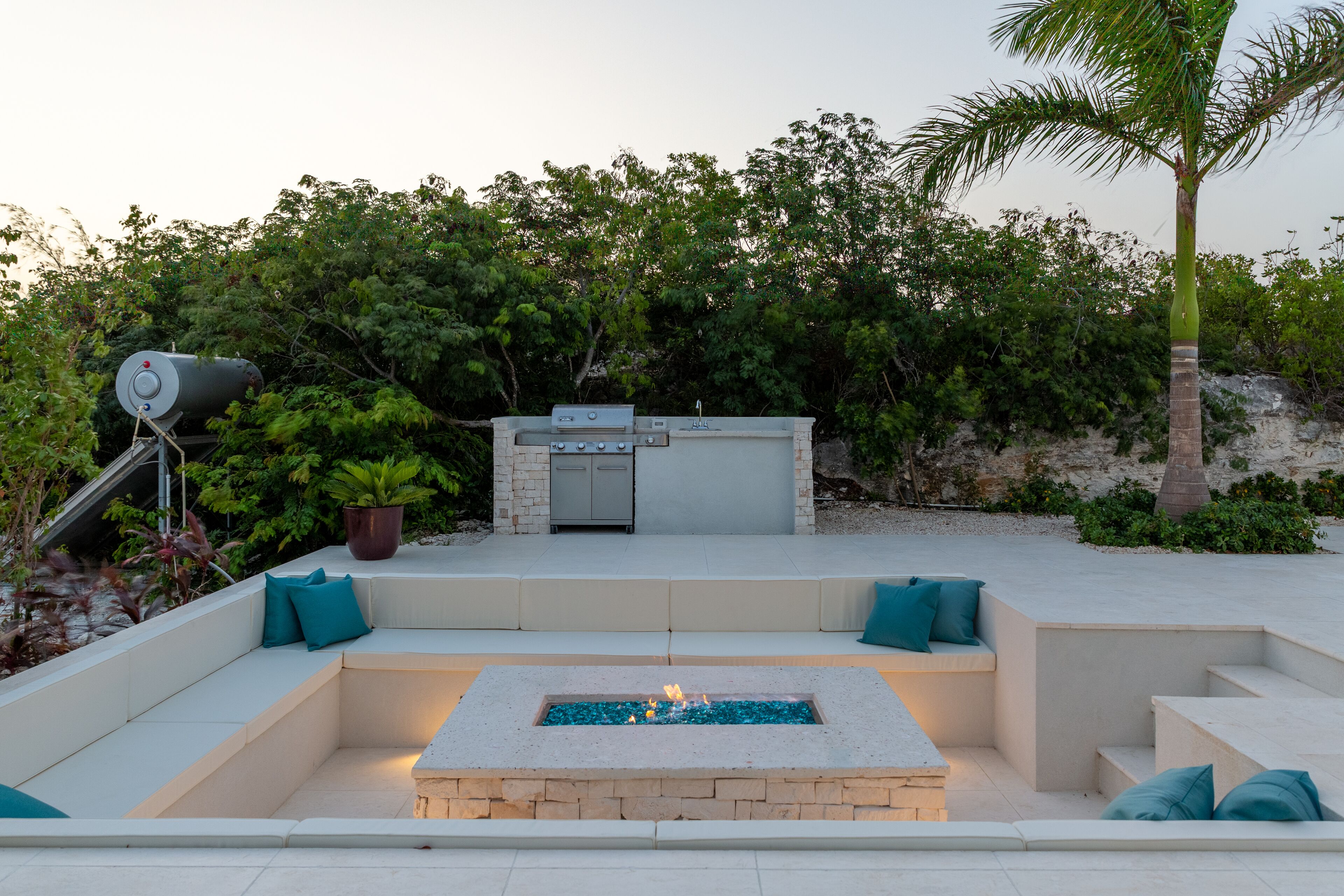 A sunken lounge with a fire pit, a grill, and an outdoor sink at dusk.