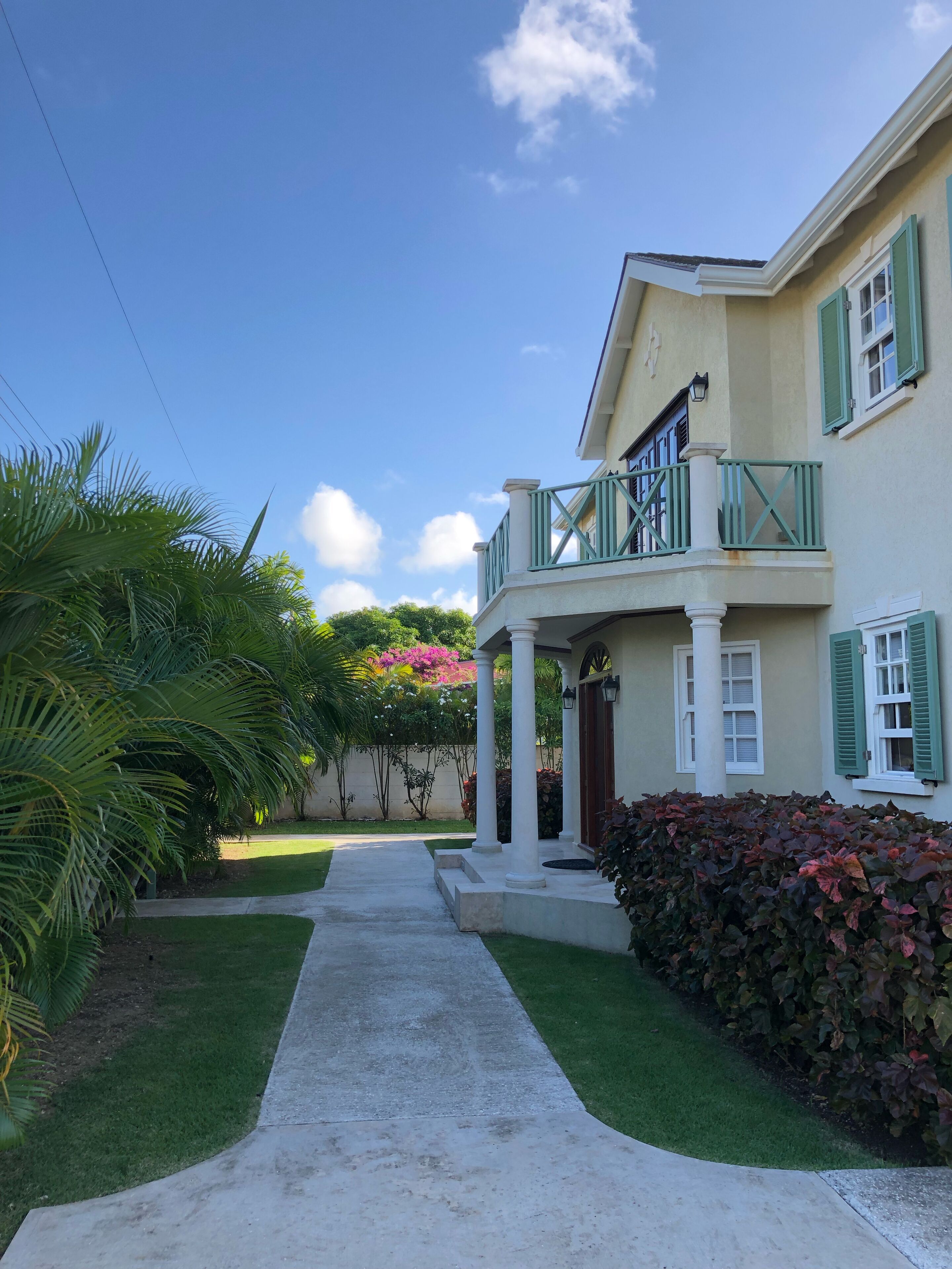 The front entrance of the villa, with a concrete walkway, palms, and a balcony above the porch.