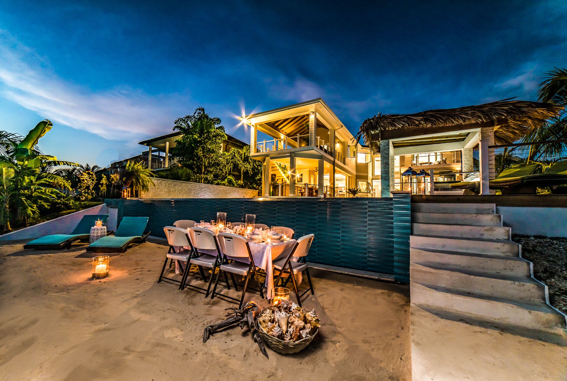 The villa and a dinner table on the sand at dusk.