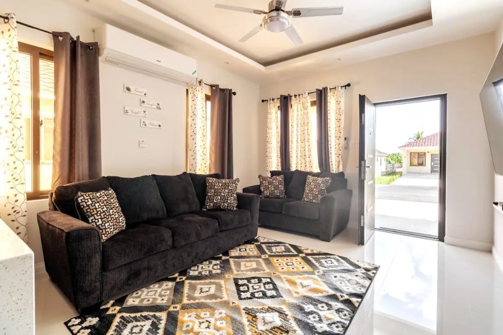 A living room with two dark sofas, a ceiling fan, and an open door to the driveway.