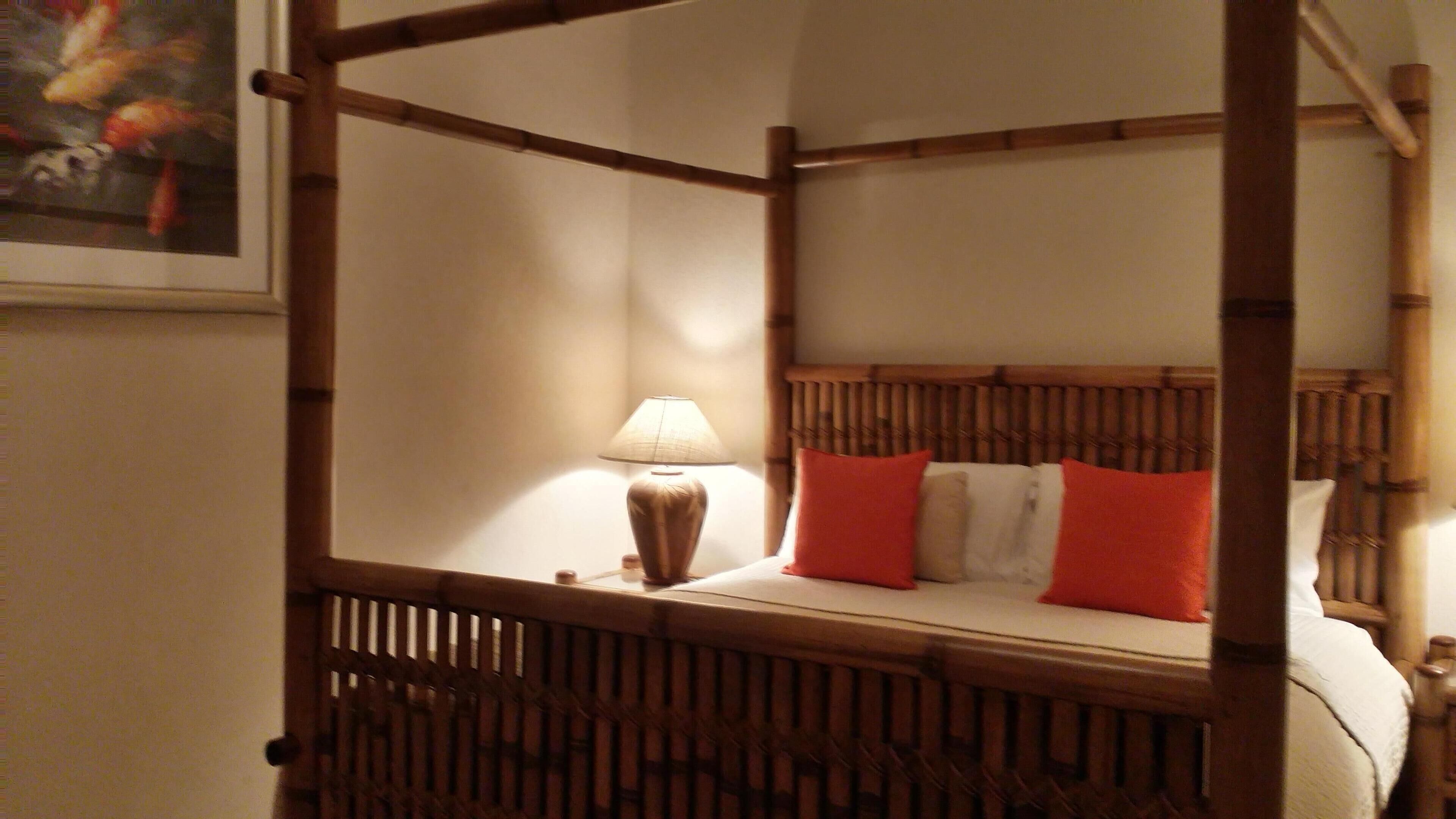A bamboo four-poster bed with orange pillows and a lit lamp.