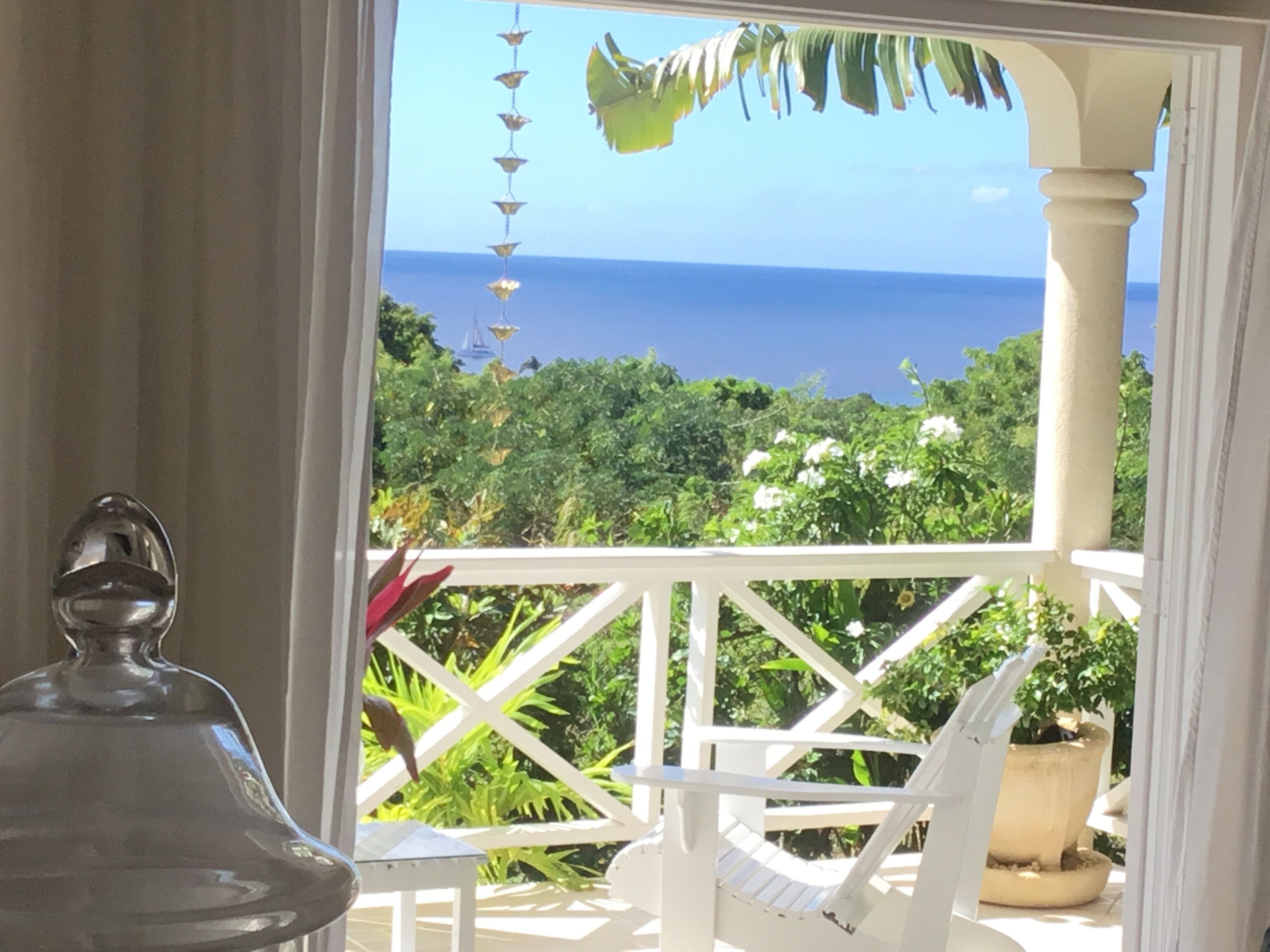A balcony with a white chair, a small table, tropical plants, and the sea beyond.