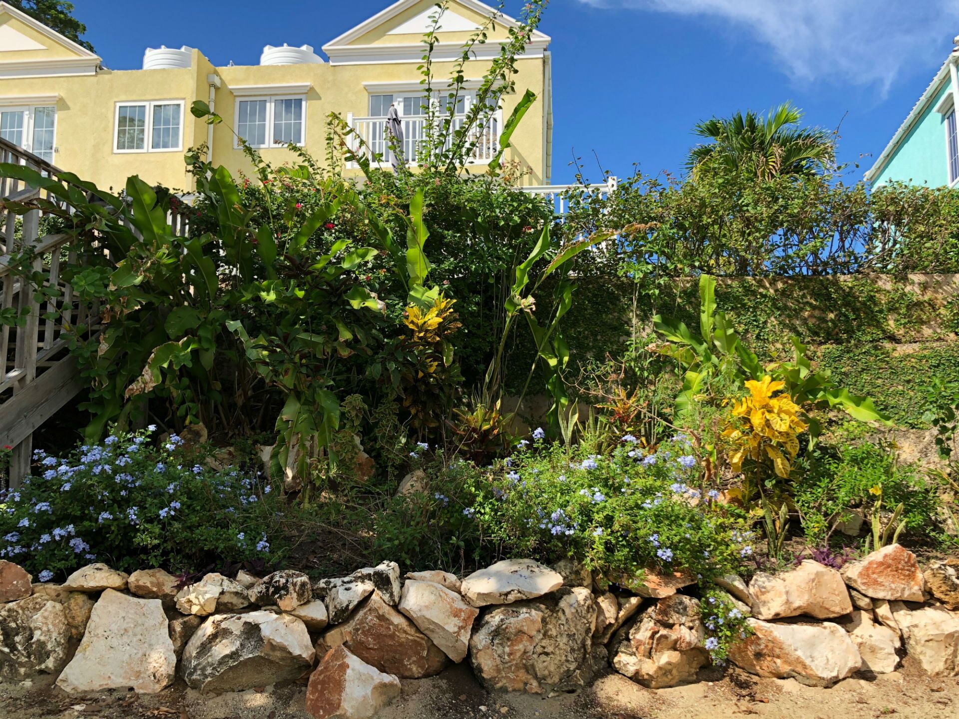 A garden with flowers and a rock wall stands in front of the yellow townhome.