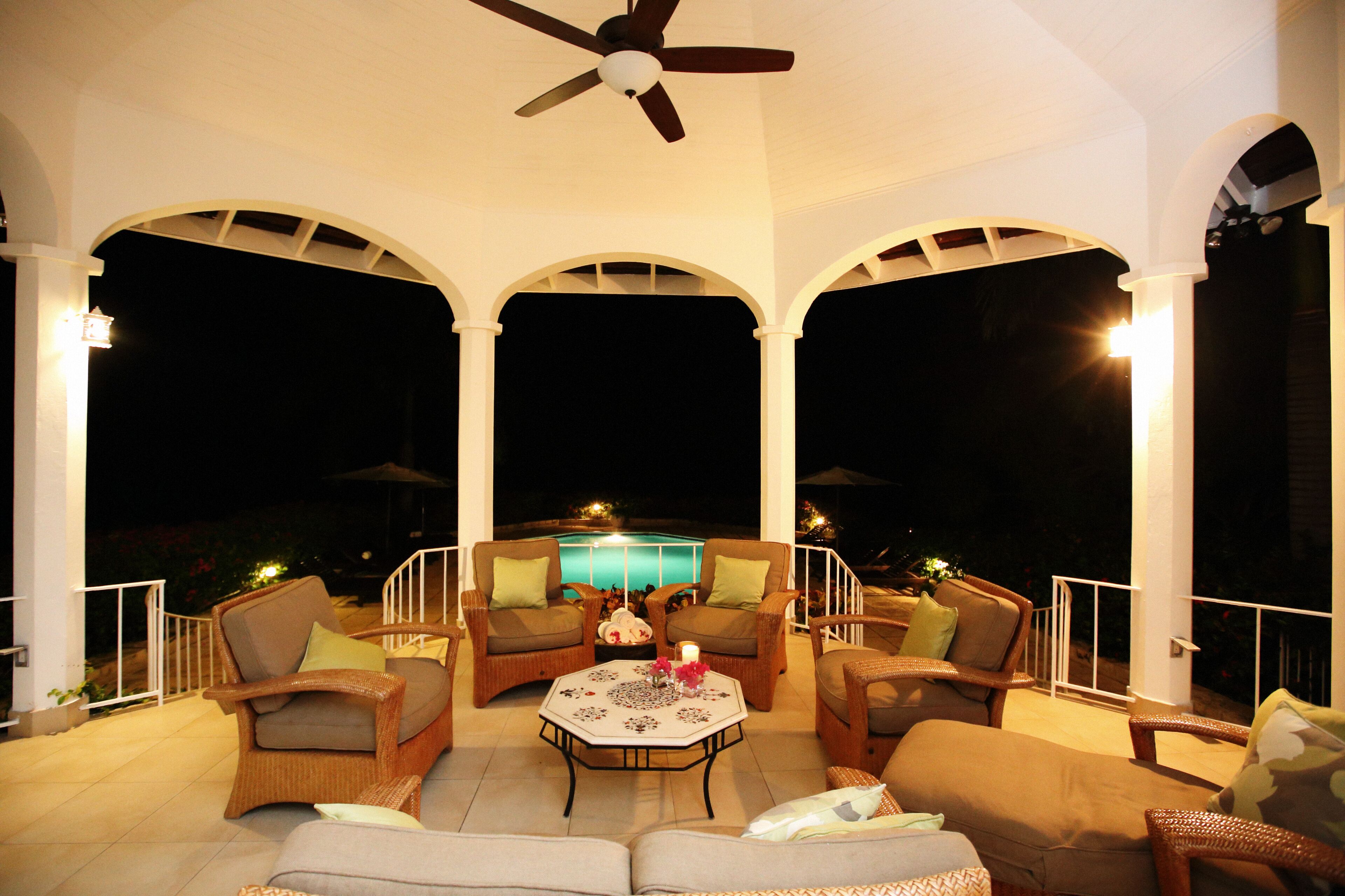 The covered veranda at night with a ceiling fan and a view of the lit pool.