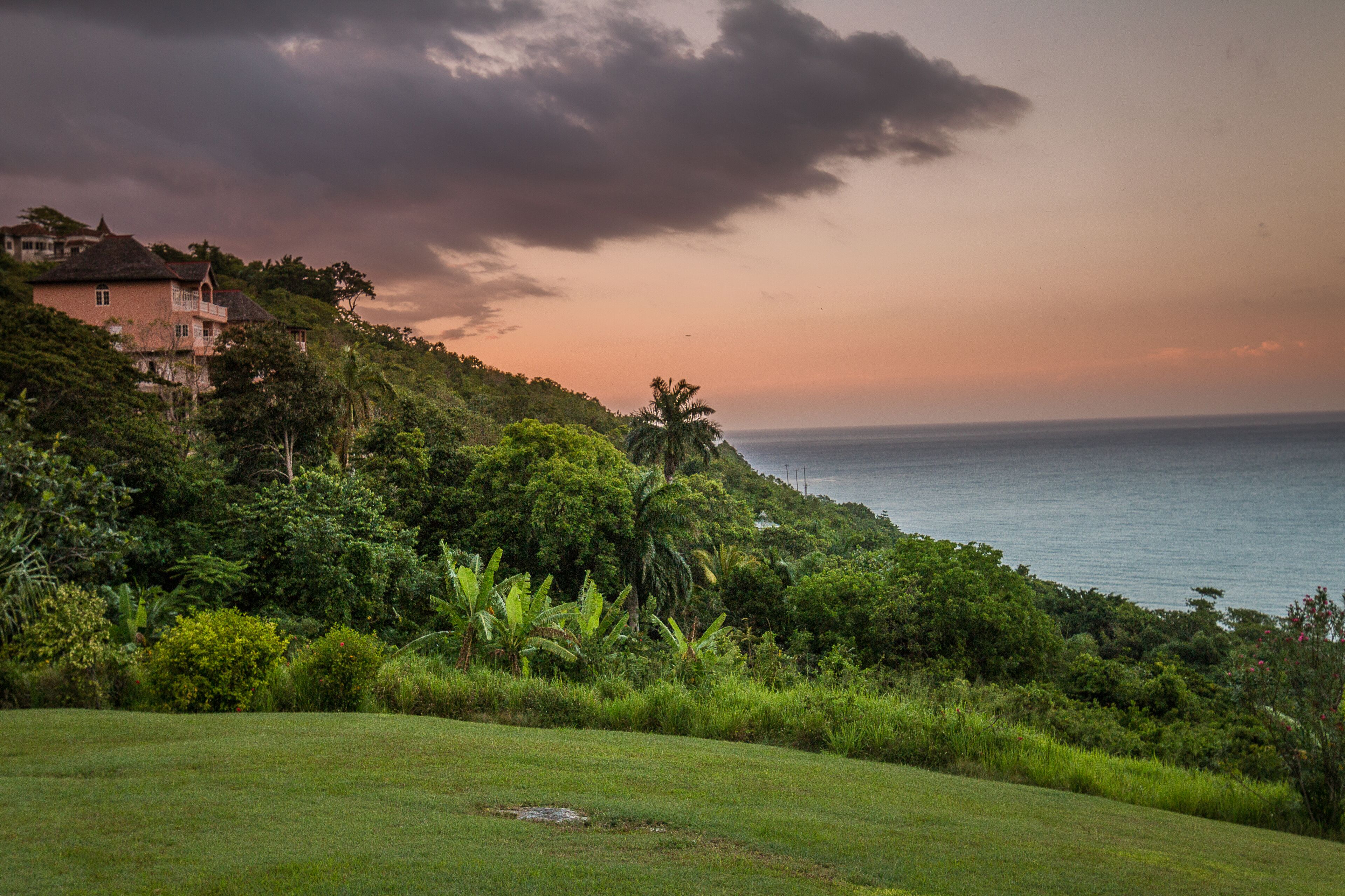 The villa stands on a green hillside above the sea at sunset.