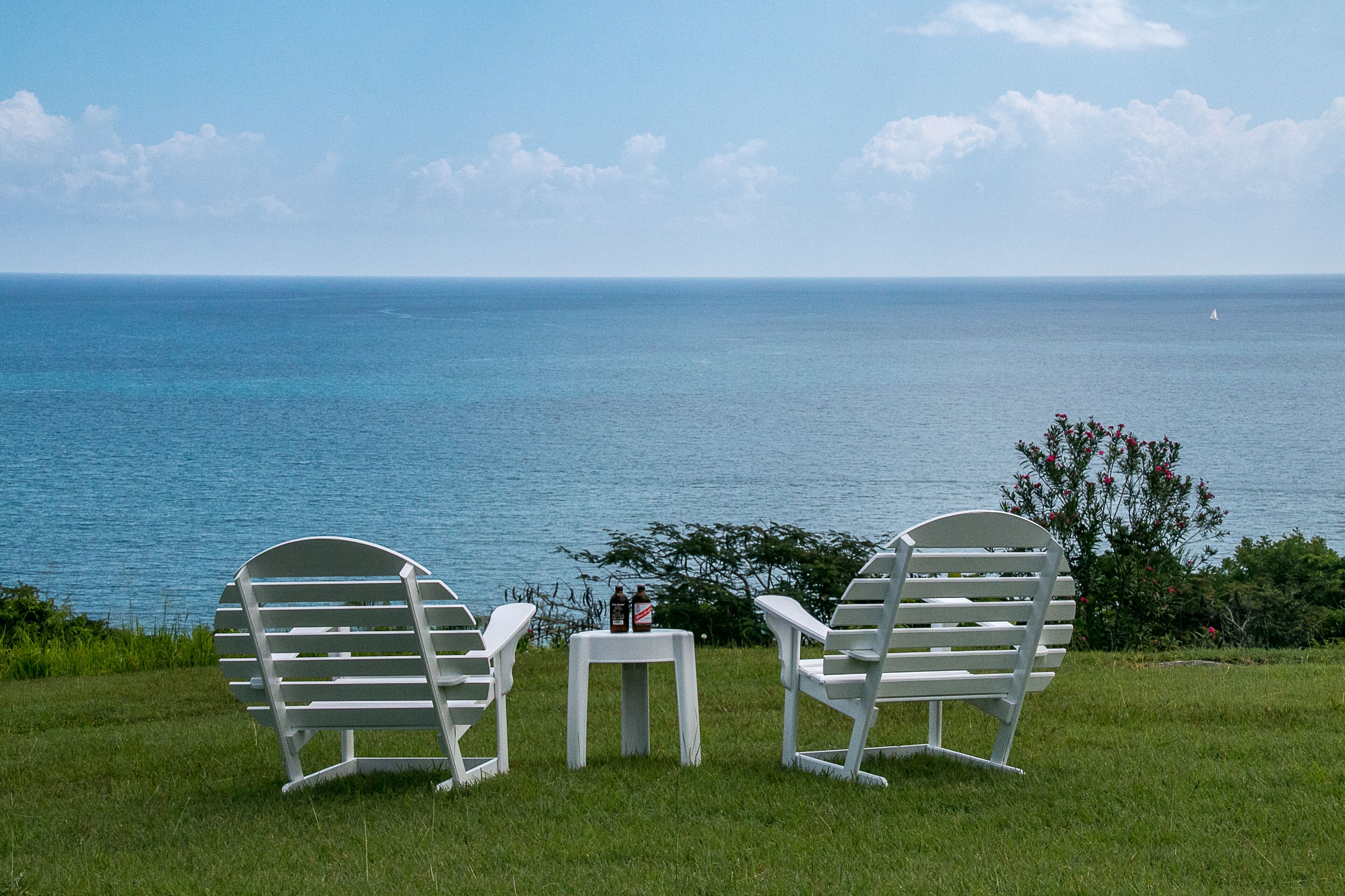 Two white chairs and a small table on the lawn face the sea.