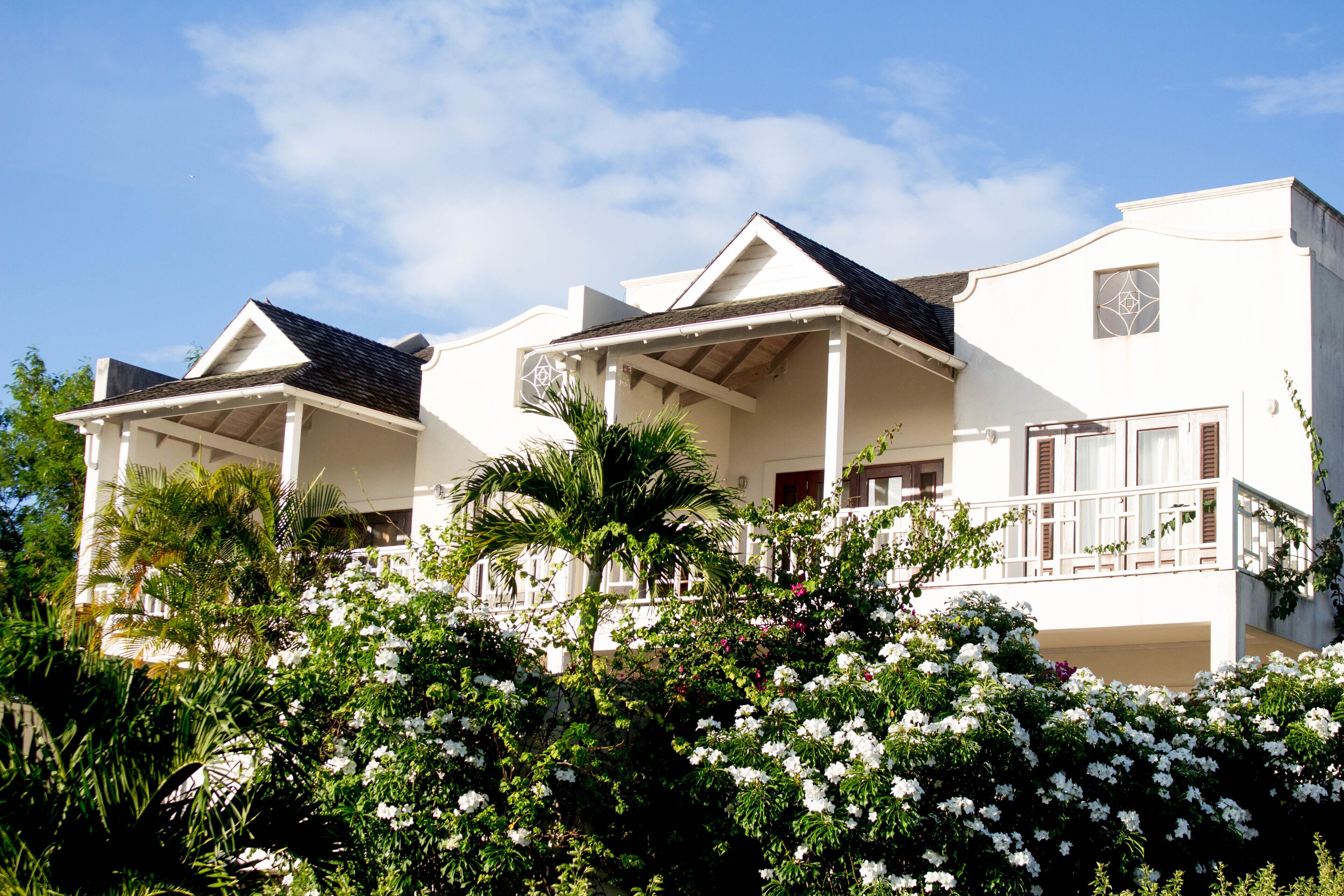 A white two-story villa with balconies and shrubs with white flowers.
