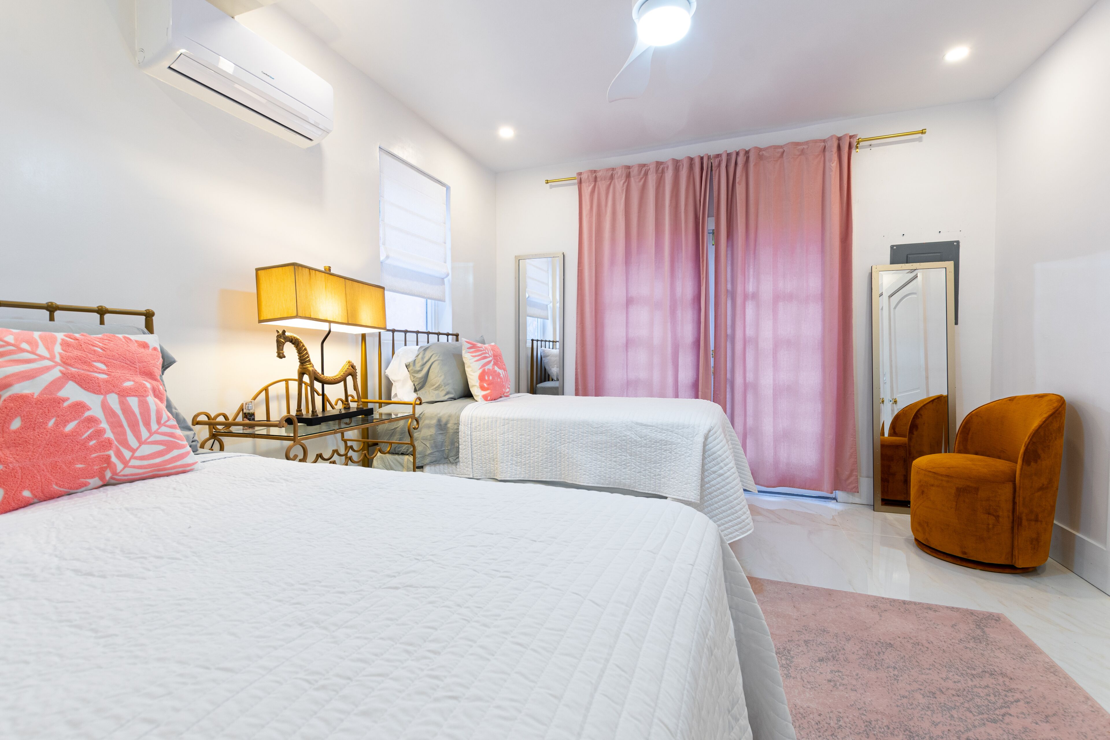 A bedroom with two beds, pink curtains, and an orange chair.