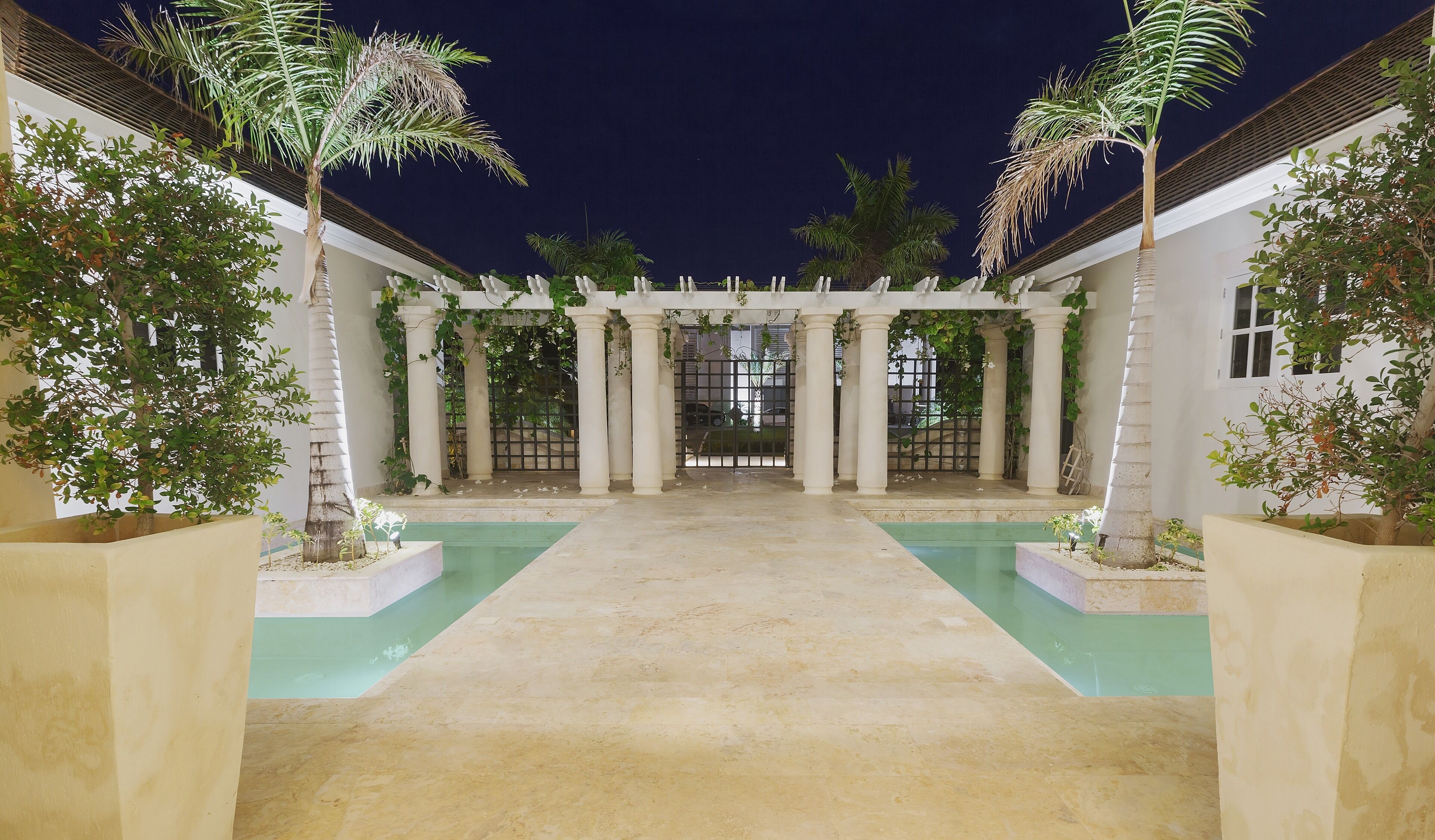 The villa courtyard at night with columns, palm trees, and two water features.