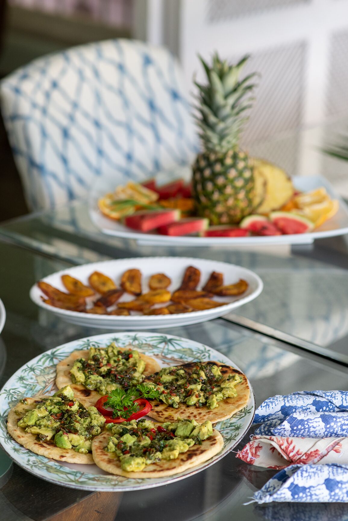 Plates of flatbread, plantain slices, and fruit on a glass table.