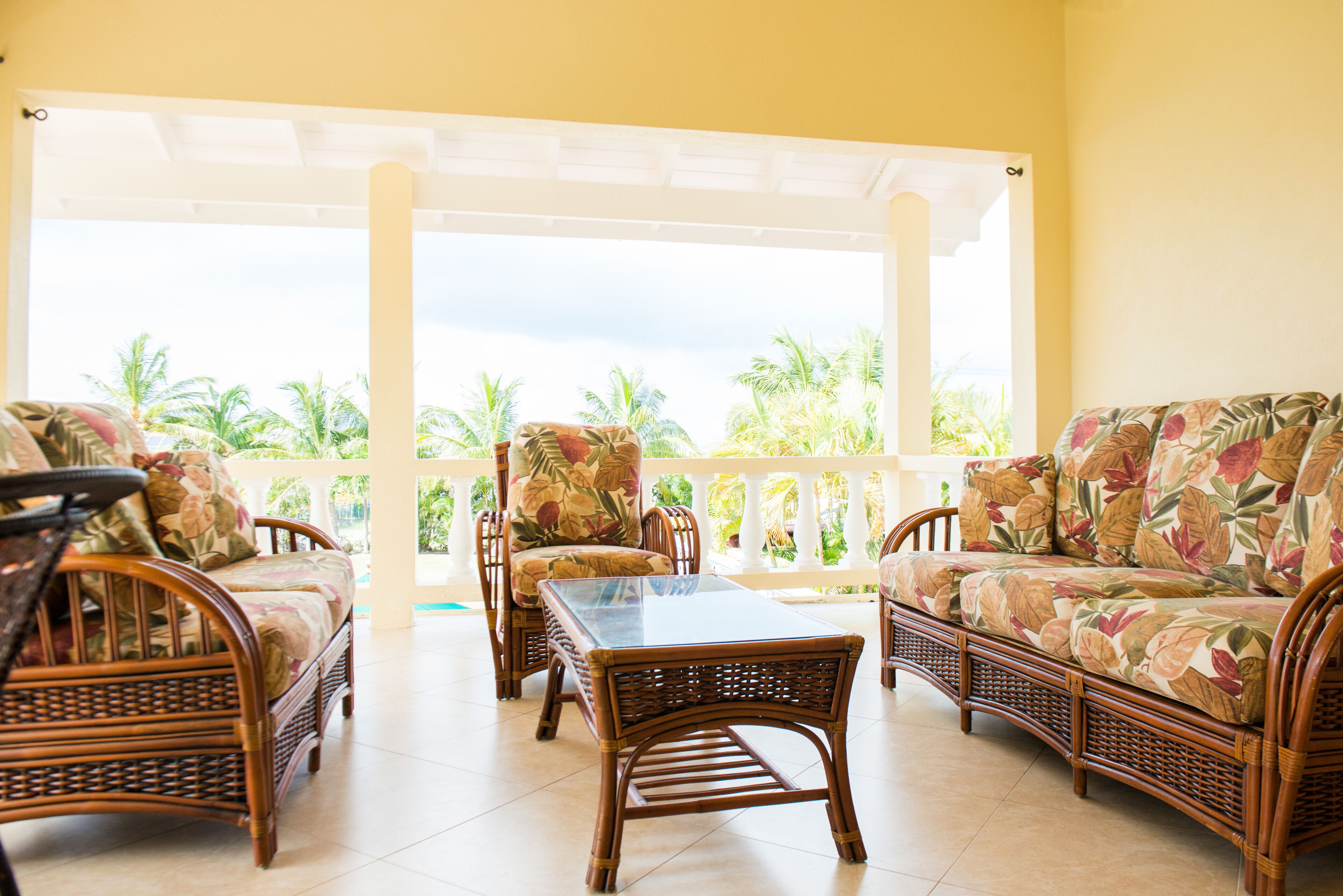 The covered veranda has rattan seats, a rattan table, and a view of palm trees.