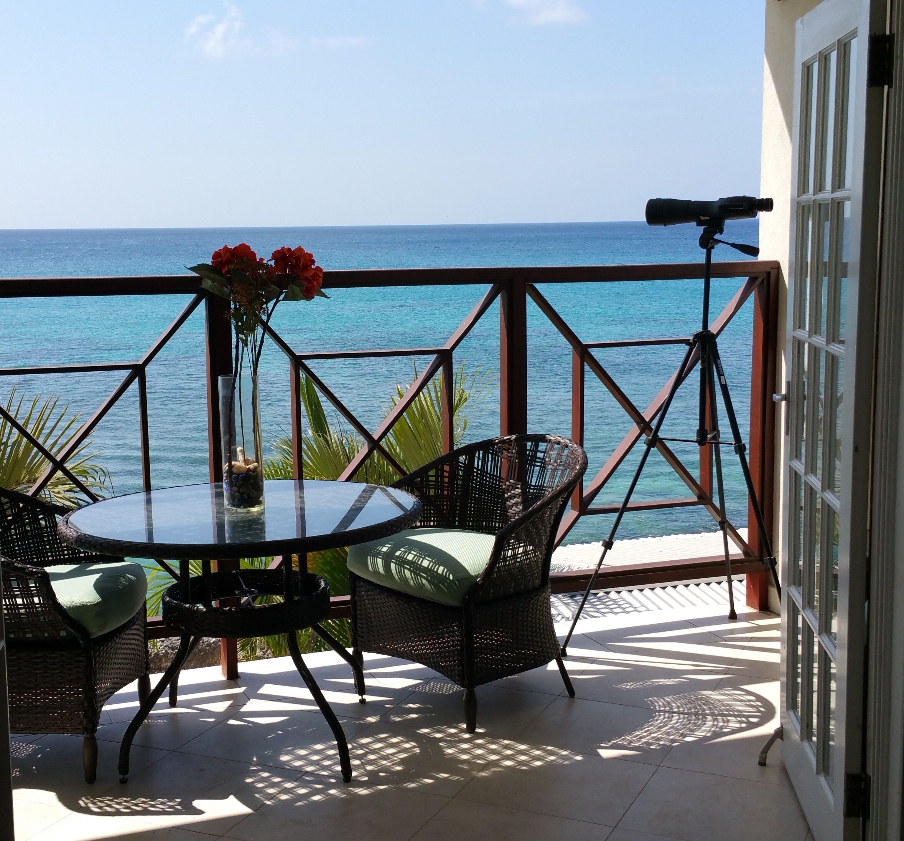 The balcony with a table, two chairs, and a spotting scope, with a view of the sea.