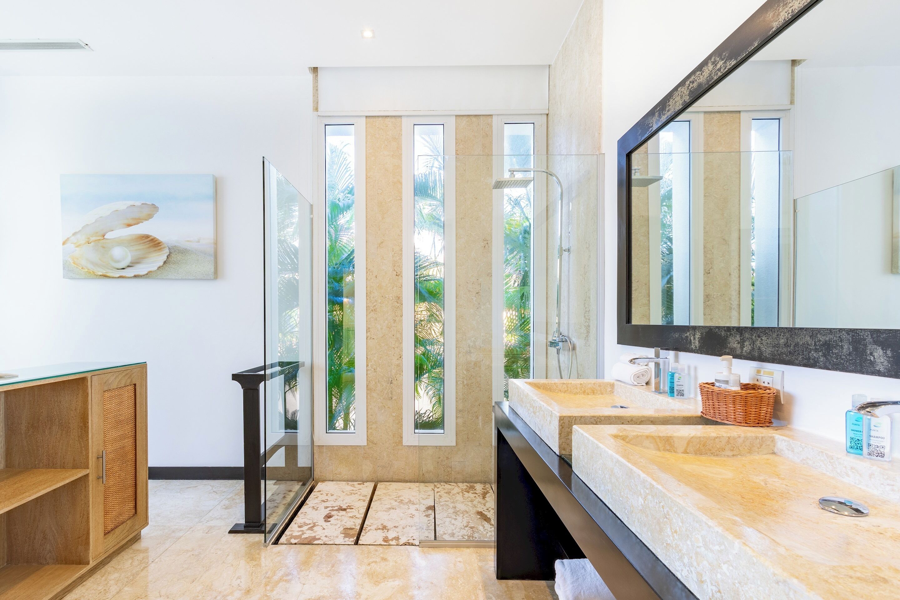 The bathroom has a walk-in shower, two stone sinks, and tall windows with a view of palm trees.