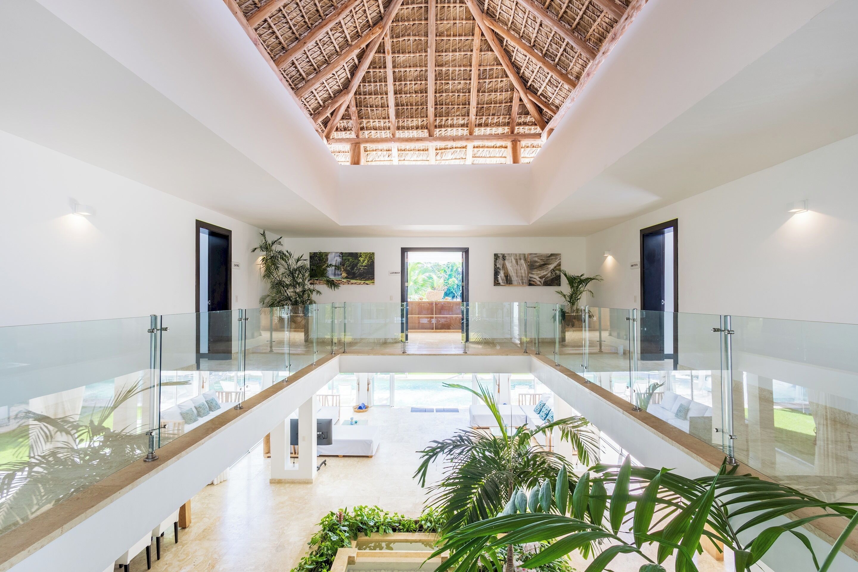 The upper landing has glass railings below a thatched roof, with a view down to the living room.