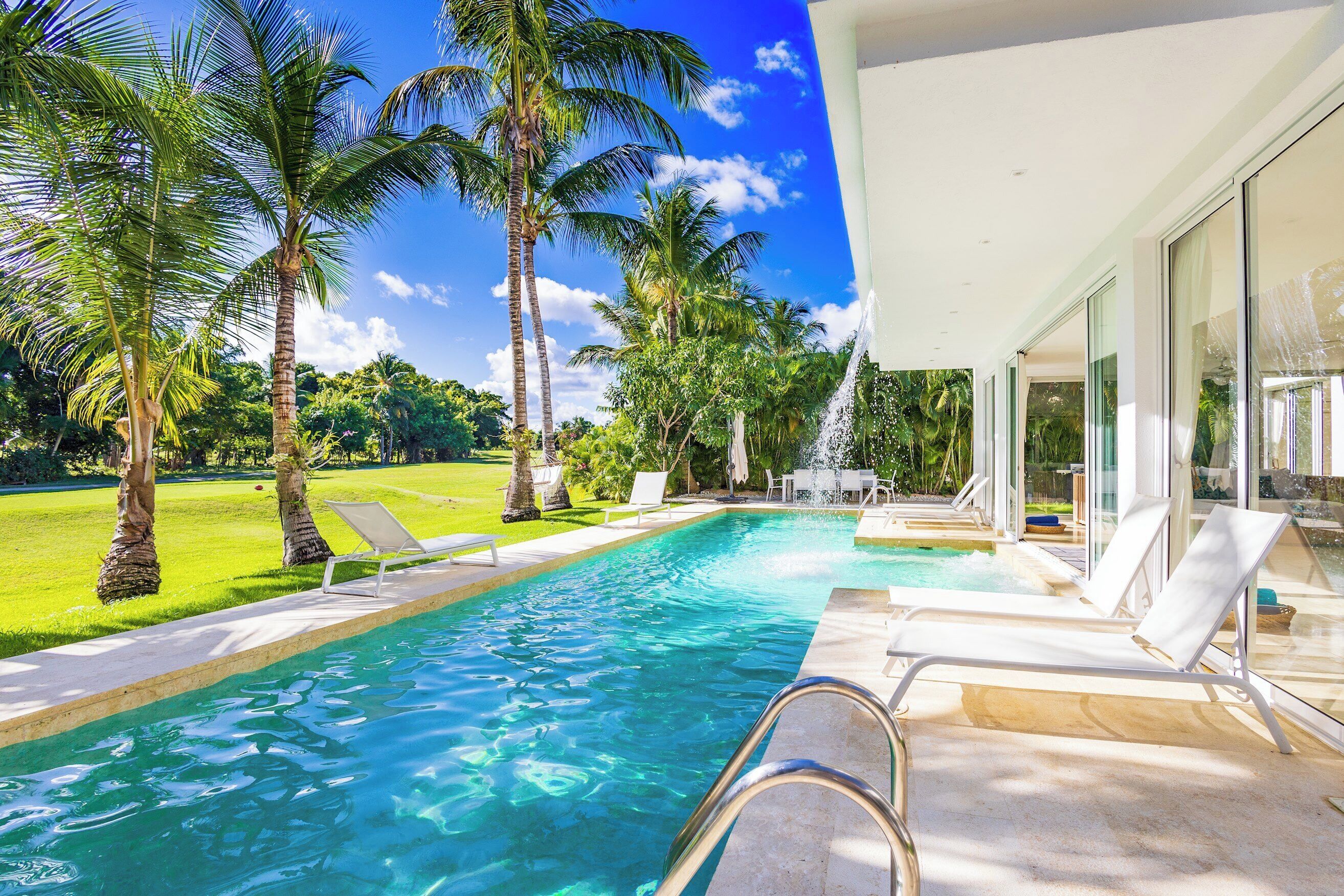 A waterfall pours from the roof into the pool beside white sun loungers and palm trees.