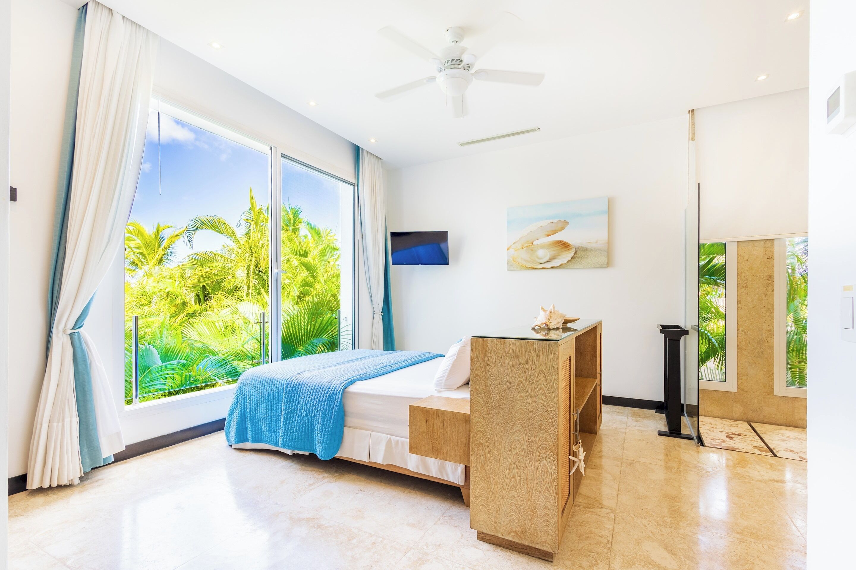The bedroom has a bed with a blue cover, a ceiling fan, and a large window with palm trees outside.