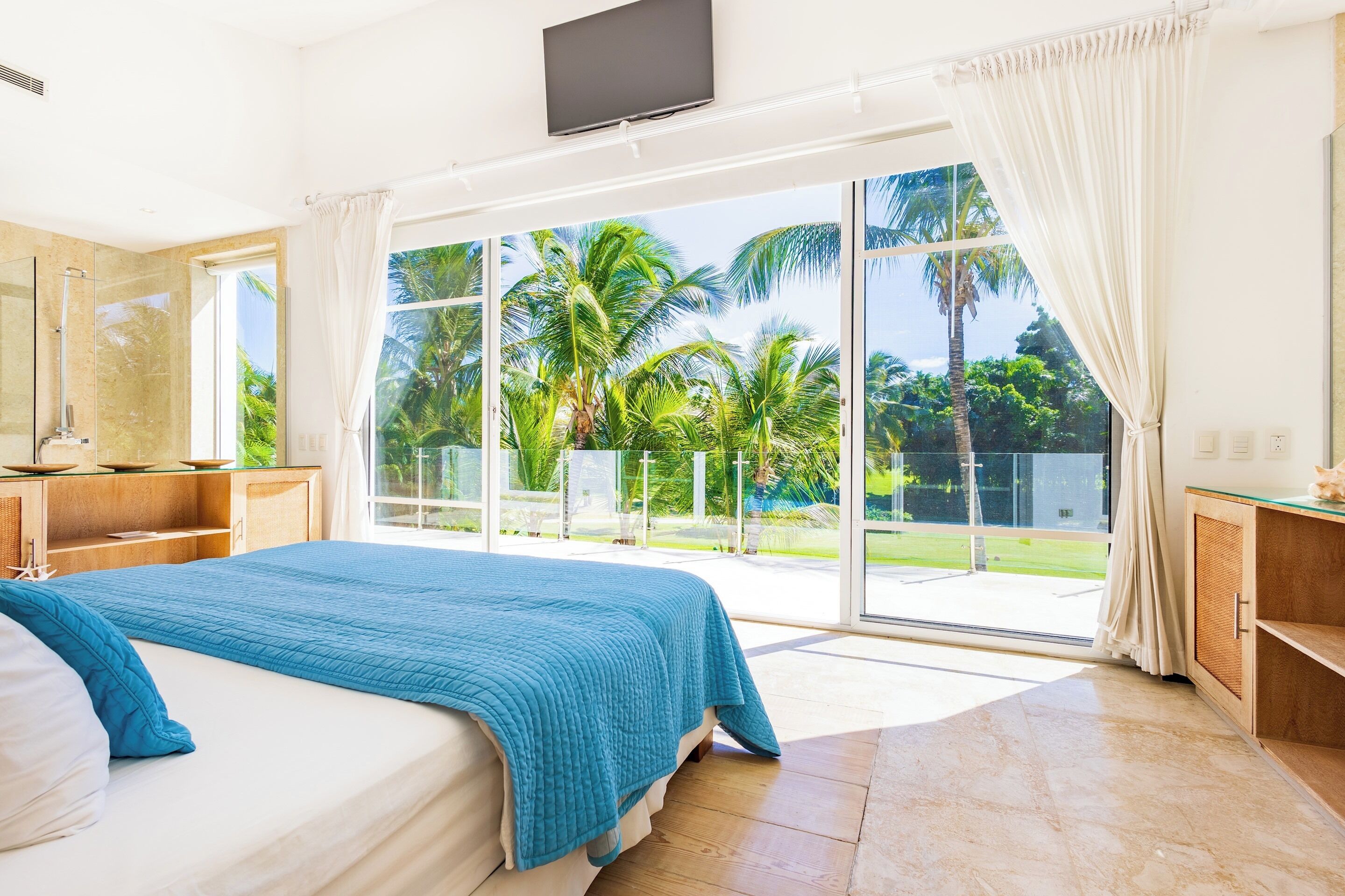The bedroom opens through glass doors to a terrace with a view of palm trees.