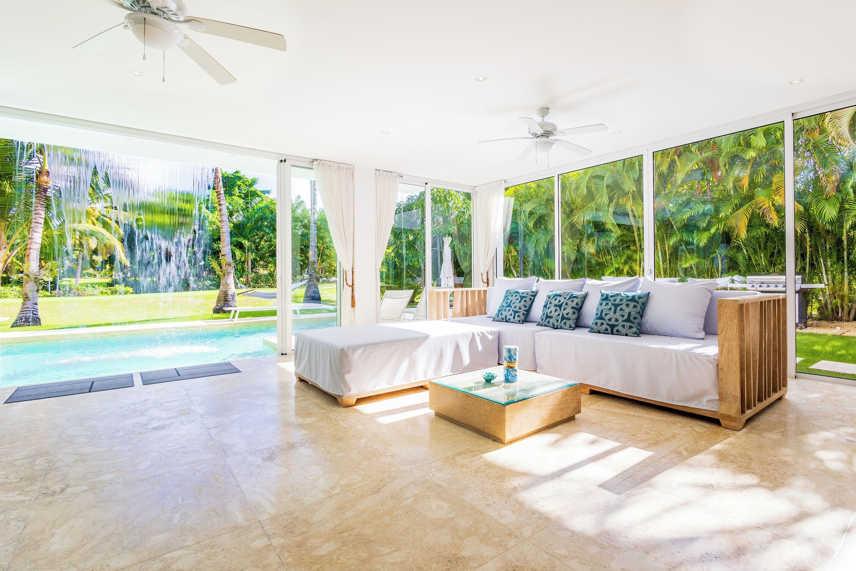 The living room has white sofas, glass walls, and a view of the pool waterfall.