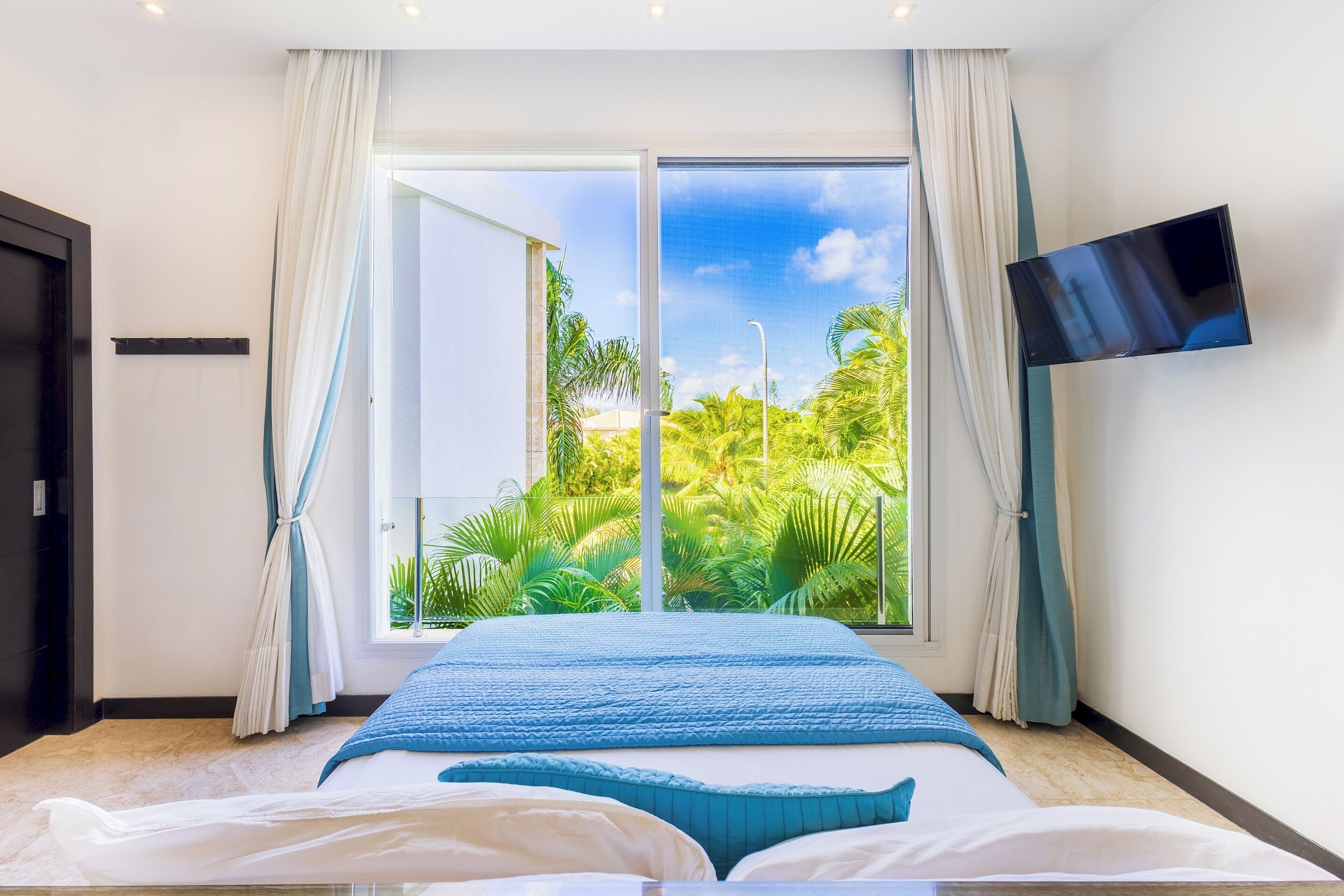 The bedroom has a bed with a blue cover, a wall television, and a window with a garden view.